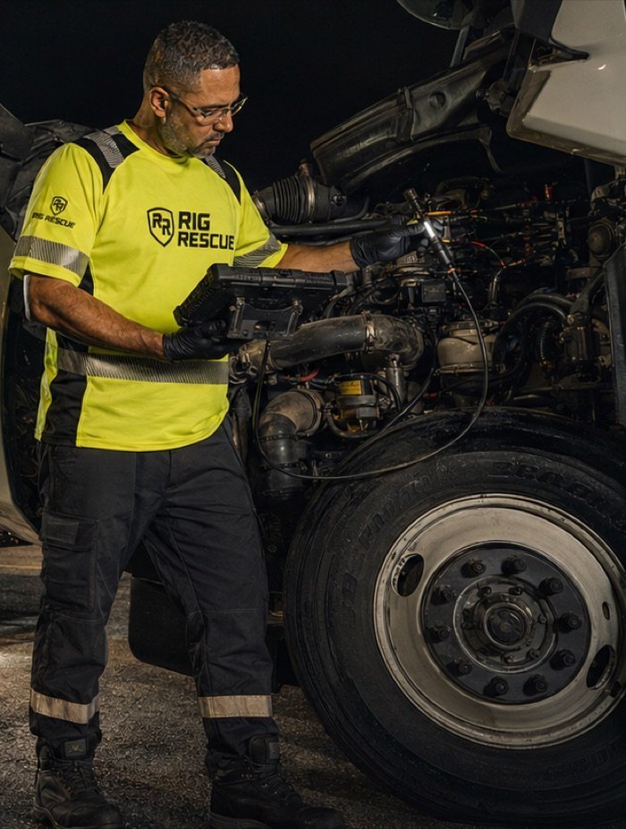 A technician in hi-vis reads a diagnostic tool at the open engine bay of a heavy truck at night.